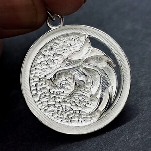 May include: A silver pendant featuring a detailed, stylized wolf head design. The pendant is circular and has a textured surface. The wolf head is cut out of the center of the pendant, creating a negative space effect.