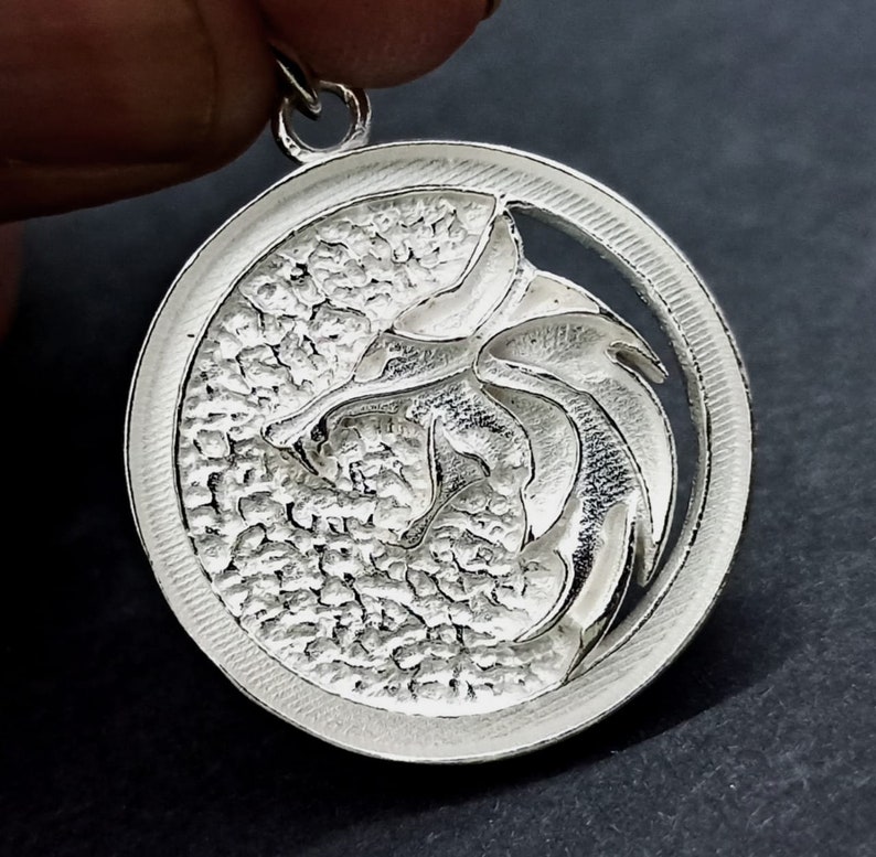 May include: A silver pendant featuring a detailed, stylized wolf head design. The pendant is circular and has a textured surface. The wolf head is cut out of the center of the pendant, creating a negative space effect.