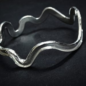 May include: A silver-colored bangle bracelet with a wavy, undulating design. The bracelet has a smooth, polished finish and reflects light, creating highlights and shadows. The text "Thevelvetsilver" is visible on the bracelet. The bracelet is displayed on a dark surface.