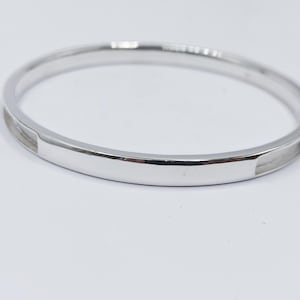 May include: A silver bangle bracelet. The bracelet has a smooth, polished finish and a rectangular design element. The bracelet is circular and appears to be made of metal. The background is white.