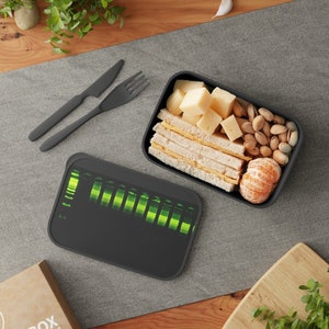 Puede incluir: Una fiambrera negra con un diseño gráfico verde y negro. La fiambrera está abierta y contiene un sándwich, queso, frutos secos y una mandarina.