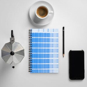 May include: A white coffee cup with a black handle sits on a white saucer. A black pencil, a silver coffee pot, and a blue and white spiral bound notebook are on a white surface.