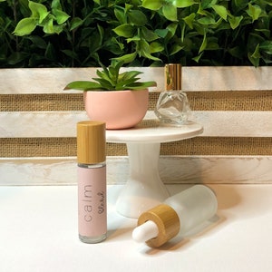 May include: Two glass bottles with bamboo lids and labels. The bottle on the left has a pink label with the text "calm blend". The bottle on the right is frosted glass. Both bottles are on a white surface.