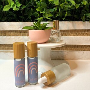 以下が含まれることがあります： 竹製の蓋と「beau」と「real」のラベルが貼られた虹のデザインの小さなガラス瓶が2本。瓶は白い表面に置かれ、小さな白いケーキスタンドと金色の蓋の透明なガラス瓶が置かれている。