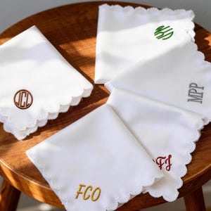 Puede incluir: Varios pañuelos blancos con bordes festoneados. Cada pañuelo está bordado con iniciales de diferentes colores: marrón, verde, gris y rojo. Los pañuelos están colocados sobre una superficie de madera.