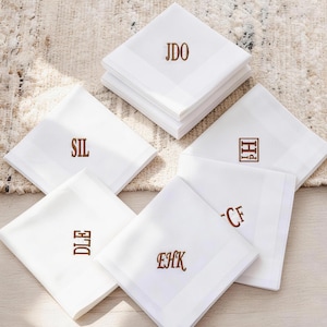 Op de afbeelding: Witte zakdoeken met bruin geborduurde initialen. Sommige zijn opgestapeld, andere uitgespreid. Elke zakdoek heeft verschillende initialen. Het ontwerp omvat een eenvoudige rand.