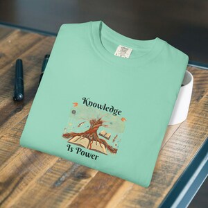 Può includere: T-shirt verde chiaro con la scritta "Knowledge Is Power" e una grafica di un albero che cresce da un libro aperto. Il design presenta elementi marroni, arancioni e verdi. La maglietta è piegata su una superficie di legno.