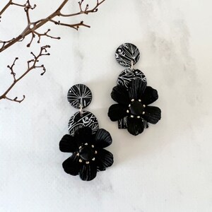 May include: Black and white floral dangle earrings with a black flower charm. The earrings are made of polymer clay and have a white floral pattern.