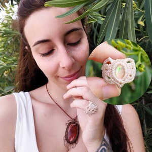 Può includere: Un anello in macramé bianco con una pietra di opale iridescente. L'anello è mostrato sul dito di una persona.