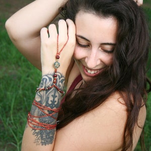 May include: A woman with long brown hair is wearing a red tank top and several bracelets on her left wrist. One bracelet is a finger ring with a red cord and a small charm. The woman has a tattoo on her left arm that says "VI-V-MMIII".