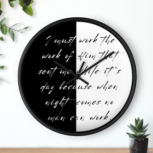 May include: Black and white clock with a black frame. The clock face is split in half, with the top half white and the bottom half black. The clock face has a quote in black text on the white half and white text on the black half: "I must work the work of Him that sent me while it's day because when night comes no man can work."