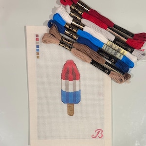 May include: A cross-stitch design of a red, white, and blue ice lolly. The canvas is next to a selection of embroidery threads in matching colours. A cursive letter "B" is in the lower right corner.