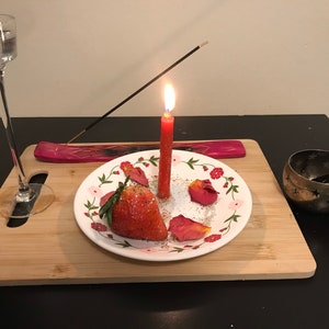May include: A red candle with a flame burning on top, a wooden incense stick, a glass of water, a plate with a strawberry, rose petals, and white powder, and a metal incense burner with a wooden spoon on a wooden cutting board.