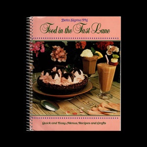 May include: A pink cookbook with a spiral binding titled "Food in the Fast Lane" by Beta Sigma Phi. The cover features a chocolate dessert with whipped cream and chocolate sauce, and two glasses of milkshakes with straws.
