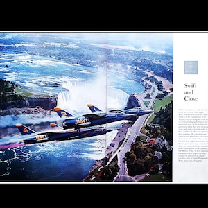 May include: An aerial view of two Navy jets flying over Niagara Falls. The jets are dark blue with orange and yellow accents. The text "NAVY" is visible on the jets. The falls are in the background.