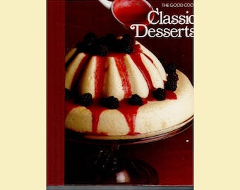 May include: A cookbook titled Classic Desserts featuring a fluted cake topped with fresh blackberries and a vibrant red berry sauce. A small white pitcher pours more red sauce over the cake, which sits on a clear glass pedestal cake stand.