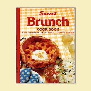 May include: A cookbook titled Sunset Brunch Cook Book with a cover image of a large baked pancake topped with fruit and cream in a skillet, a glass of juice, and a small jar of jam on a yellow and white checkered tablecloth background.