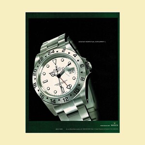 May include: A silver Rolex Oyster Perpetual Explorer II wristwatch with a white dial, black hour markers, and a 24-hour bezel. The watch features a date window with a cyclops lens, a stainless steel bracelet, and the text Oyster Perpetual Explorer II.