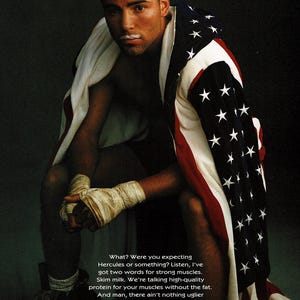 1996 magazine ad featuring boxer Oscar De La Hoya with boxing wraps, an American flag draped over his shoulders, and a milk mustache as part of the Got Milk? campaign. Dark background with bold text promoting skim milk for strong muscles. Original single page about 8×10 with normal vintage wear.