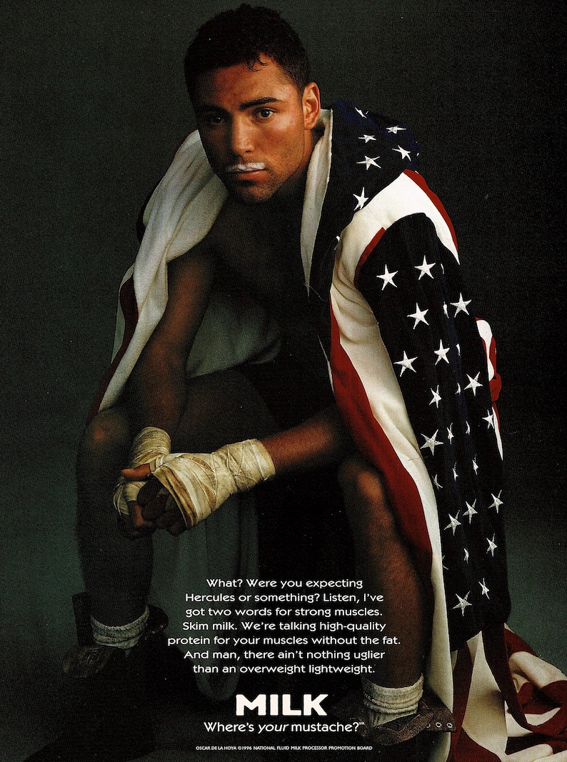 1996 magazine ad featuring boxer Oscar De La Hoya with boxing wraps, an American flag draped over his shoulders, and a milk mustache as part of the Got Milk? campaign. Dark background with bold text promoting skim milk for strong muscles. Original single page about 8×10 with normal vintage wear.