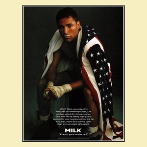 1996 magazine ad featuring boxer Oscar De La Hoya with boxing wraps, an American flag draped over his shoulders, and a milk mustache as part of the Got Milk? campaign. Dark background with bold text promoting skim milk for strong muscles. Original single page about 8×10 with normal vintage wear.
