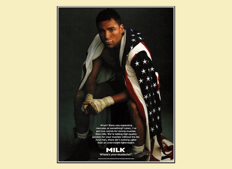 1996 magazine ad featuring boxer Oscar De La Hoya with boxing wraps, an American flag draped over his shoulders, and a milk mustache as part of the Got Milk? campaign. Dark background with bold text promoting skim milk for strong muscles. Original single page about 8×10 with normal vintage wear.