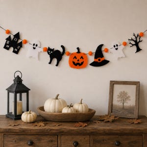 Op de afbeelding: Een Halloween-slinger met viltuitsparingen van een zwart spookhuis, witte spoken, een zwarte kat, een oranje pompoen, een zwarte heksenhoed en een zwarte boom, verbonden door kleine oranje vilten balletjes aan een dun zwart koord.
