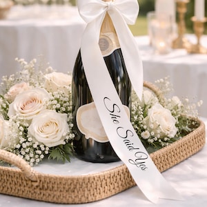 Puede incluir: Una botella de champán con una cinta blanca con el texto "She Said Yes". La botella está rodeada de rosas blancas y rosadas y paniculata, presentada en una bandeja tejida, ideal para una celebración.