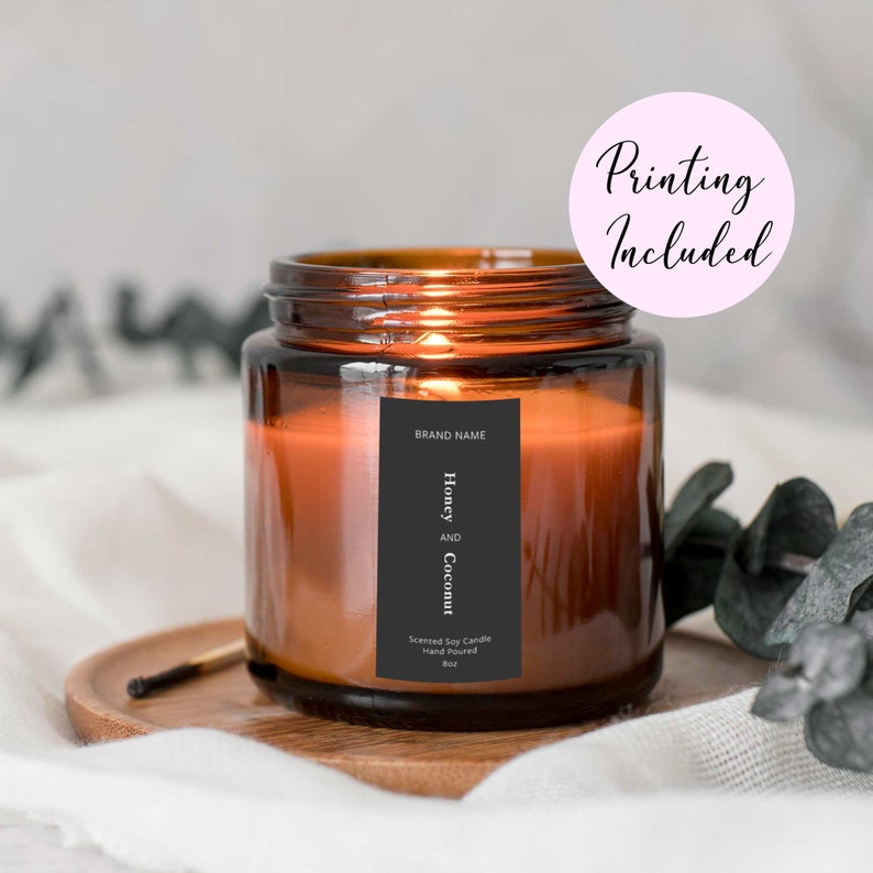 Op de afbeelding: Een bruine glazen kaars pot met een zwart label waarop staat "Honey and Coconut" en "Scented Soy Candle Hand Poured 8oz". De kaars is aangestoken en het label bevat ook de tekst "Brand Name".
