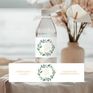 Peut inclure: Étiquette de bouteille d'eau avec une couronne de feuilles vertes et des initiales dorées "D & E" avec le texte "Drake & Elina 10.09.20XX" et "Merci de célébrer avec nous !"