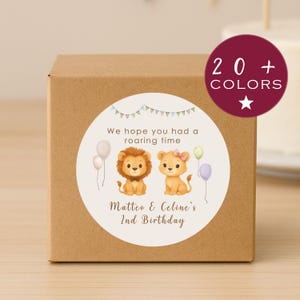 May include: A brown cardboard box with a round sticker featuring two cartoon lions, balloons, and the text "We hope you had a roaring time." The sticker also includes the text "Matteo & Celine's 2nd Birthday." A burgundy circle with the text "20+ COLOURS" is in the upper right.