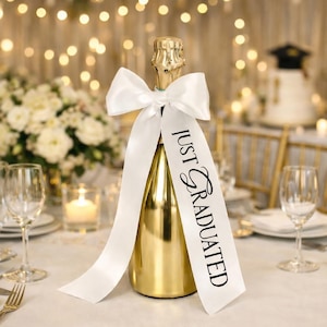 May include: A gold champagne bottle with a white ribbon and the text "JUST GRADUATED". The bottle is on a table set for a celebration, with glasses, flowers, and a cake in the background. The ribbon is tied in a bow.
