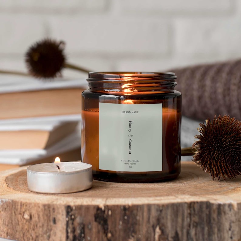 Op de afbeelding: Een bruine glazen kaars pot met een wit label met de tekst "BRAND NAME Honey AND Coconut Scented Soy Candle Hand Poured 8oz". De kaars is aangestoken en er staat een kleine zilveren kaarsenhouder met een brandende kaars voor.
