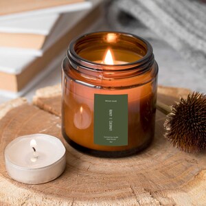 Op de afbeelding: Een bruine glazen kaars pot met een groen label waarop staat "BRAND NAME" "HONEY AND COCONUT" "Scented Soy Candle Hand Poured 8oz". De kaars is aangestoken en de vlam is zichtbaar. De kaars staat op een houten oppervlak met een kleine witte theelichtkaars en een gedroogde bruine bloem.