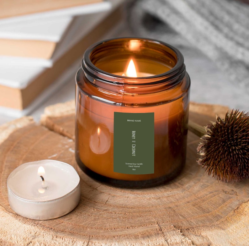 Op de afbeelding: Een bruine glazen kaars pot met een groen label waarop staat "BRAND NAME" "HONEY AND COCONUT" "Scented Soy Candle Hand Poured 8oz". De kaars is aangestoken en de vlam is zichtbaar. De kaars staat op een houten oppervlak met een kleine witte theelichtkaars en een gedroogde bruine bloem.