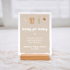 May include: A baby shower sign with the text "books for baby" and a message thanking guests for starting a baby's first library. The sign is beige with a wooden base and features baby-themed illustrations.