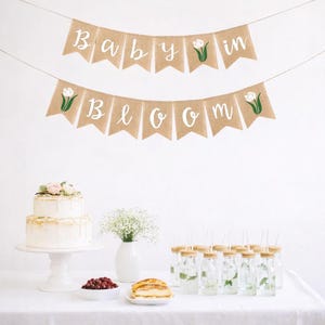 May include: A festive banner with the words "Baby in Bloom" in white script, accented with green tulip illustrations, hangs above a table set for a celebration. A two-tiered cake, drinks, and snacks complete the scene.