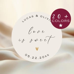 Può includere: Un adesivo rotondo bianco con il testo "Lucas & Olivia" e "Love is sweet" in scrittura corsiva nera. La data "05.22.20XX" è stampata sotto il testo. Un piccolo cuore dorato si trova sopra la data. Un cerchio bordeaux con una stella bianca e il testo "20+ COLORS" si trova nell'angolo in alto a destra dell'immagine.