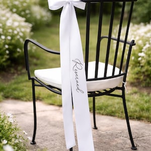 Op de afbeelding: Zwarte metalen stoel met een wit kussen en een wit lint dat aan de rugleuning is vastgemaakt. Op het lint staat het woord "Reserved" in zwart schrift. De stoel staat buiten, waarschijnlijk voor een bruiloft of evenement.