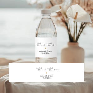 Könnte beinhalten: Ein weißes Hochzeitsetikett für eine Wasserflasche mit dem Text "Mr & Mrs" in Kursivschrift, einem kleinen Herzsymbol, den Namen "Ariana & Drake" und dem Datum "10.09.20xx".