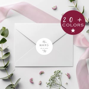 May include: A white envelope with a circular sticker reading "RSVP by" and the date "06.22.20xx" in black, with a floral design. A burgundy circle with "20 + COLORS" is in the upper right. Pink ribbon and flowers surround the envelope.