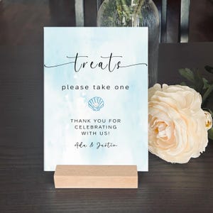 May include: A light blue sign with a watercolour background and the text "treats please take one" with a seashell graphic. The sign also includes the text "Thank you for celebrating with us! Ada & Justin".