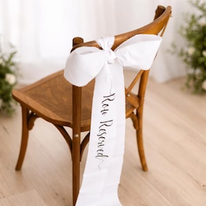 May include: A wooden chair with a white fabric bow and a long white sash. The sash is printed with the words "Row Reserved" in a delicate script. The chair has a classic design and is set in a bright, airy space.