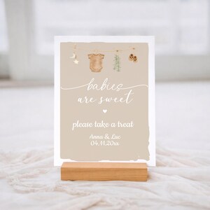 May include: A baby shower sign with the text "babies are sweet, please take a treat." The sign features illustrations of baby clothes and a wooden base. The sign is light beige with white text and a white border.