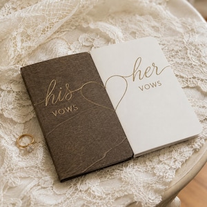 Può includere: Due libretti per i voti rettangolari, uno in legno marrone scuro e uno bianco, con scritte dorate his vows e her vows, un contorno a cuore dorato che unisce i due libretti, accompagnati da un anello dorato con una pietra trasparente.