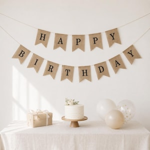 May include: A birthday party scene with a banner that reads "HAPPY BIRTHDAY". The banner is made of brown burlap material with black letters. A white cake sits on a wooden stand, next to a gift box and balloons.