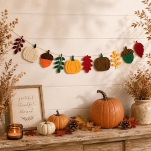 May include: A felt garland featuring alternating shapes of pumpkins, acorns, and leaves in shades of cream, orange, yellow, green, brown, and red, strung on a white cord. The garland is displayed against a white horizontal panelled wall.