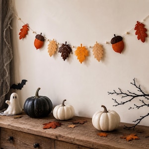 Könnte beinhalten: Eine dekorative Girlande mit gefilzten herbstlichen Blättern und Eicheln in Orange, Gelb und Braun, die auf einer Schnur mit kleinen Holzperlen aufgereiht sind. Die Girlande ist horizontal angeordnet und zeigt herbstliche Formen.
