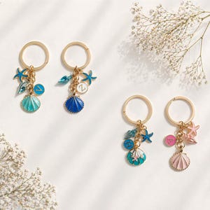 May include: Four gold keyrings with marine-themed charms. Each keyring has a gold ring and a chain with charms such as seashells, starfish, and gems in blue, pink, and turquoise. White flowers are in the background.