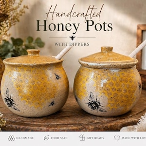 Könnte beinhalten: Zwei handgefertigte Honigtöpfe mit Dippern. Die Töpfe sind beige mit gelben Bienenwaben- und Bienenmotiven. Die Deckel sind oben und die Dipper sind innen. Der Text "Handcrafted Honey Pots" steht oben.